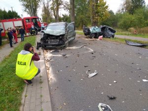 na pierwszym planie policjant zabezpieczający ślady zdarzenia na drugim planie dwa pojazdy biorące udział w wypadku drogowym