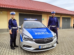 z lewej strony policjantka z prawej policjant a pośrodku radiowóz