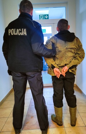 z lewej strony soi policjant tyłem, trzyma mezczyznę w kajdankach założonych na tęce trzymane z tyłu
