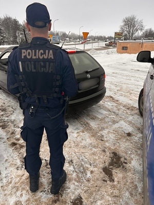 policjant stoi tyłem w tle czarny samochód i część radiowozu policyjnego